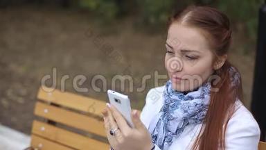 红发中年妇女，手里拿着智能手机，坐在公园的长凳上，在社交中交流
